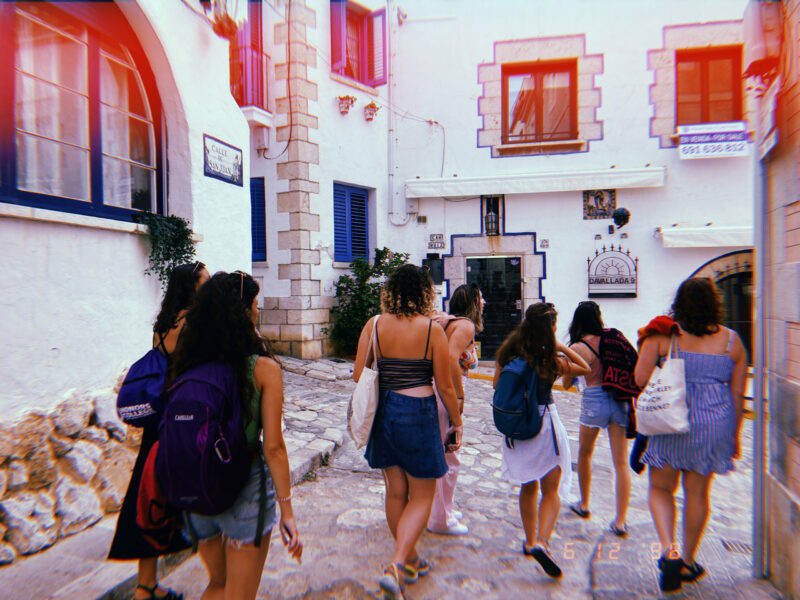 A group of girls walk around the corner at the end of an alley. The walls are a bright, white brick. The girls are dressed in summer clothes. The picture is taken so you can only see the back of each girl and not their faces.