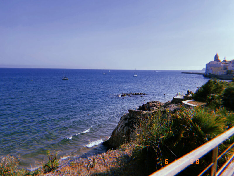 Overlooking a cliffside. On the bottom right corner there are rocks and green plants that go down the side of the hill. On the rest of the image there is bright blue water and bright blue skies.
