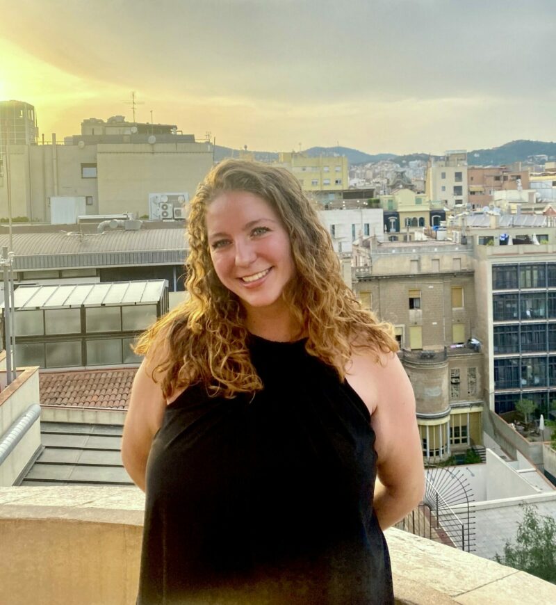A girl stands in a black dress and looks at the camera. Her hair is brown and loose, flowing down her shoulders. She smiles. In the background the city stretches on showing numerous buildings and even mountains in the very back.