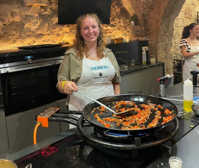 A girl stands wearing an apron and smiling. She holds a spatula, the cooking end sits in a large pans. The girl is stirring vegetables. She is in a kitchen with cozy lighting.