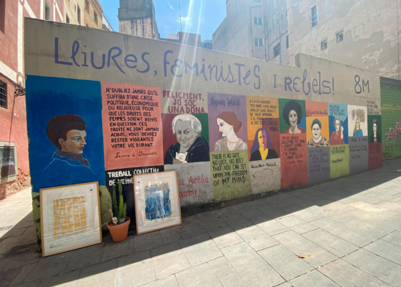 A wall is covered with a painted mural. Across the top it says "Liures, feministes, y rebels". The mural is of several quotes and several portraits. Each new image sits over a different colored background.